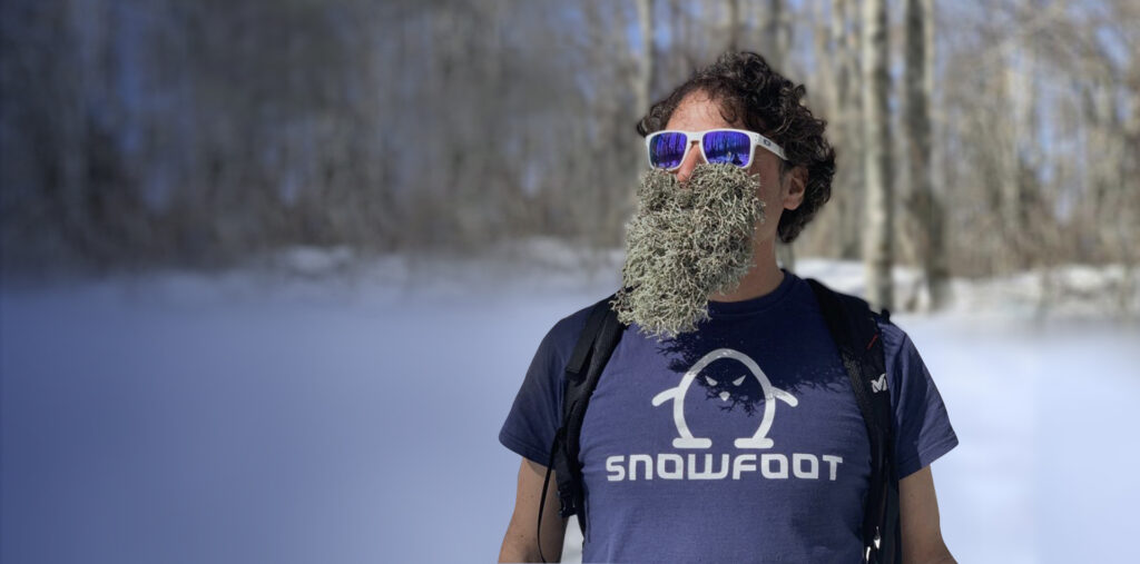 A bearded man wearing sunglasses and a Snowfoot Snowshoes shirt in the middle of a snowy forest