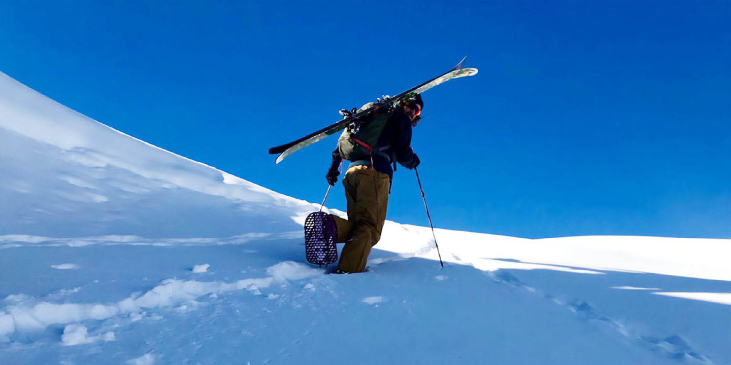 A backcountry skier snowshoeing up a mountain with skis on their back