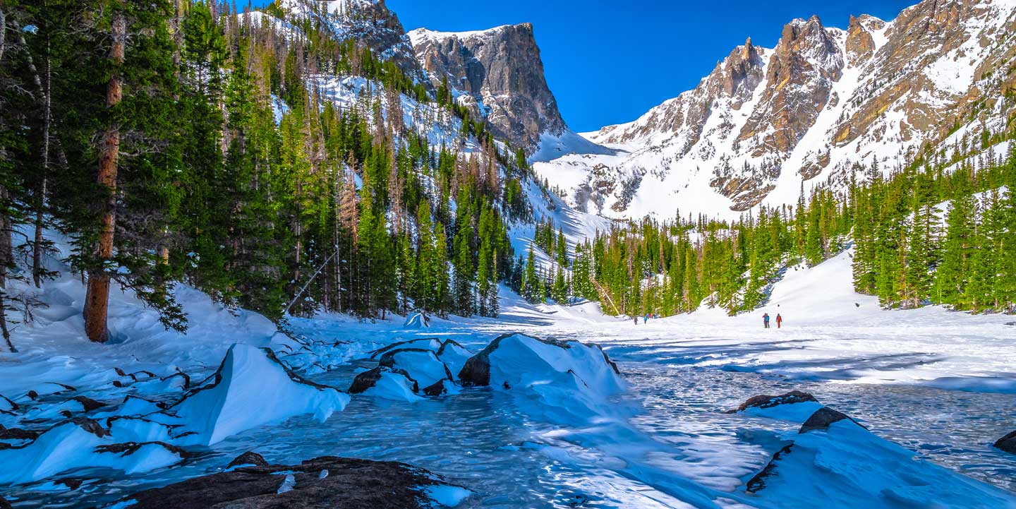 A creek running through imposing mountains with green pine trees and snowshoers in the distance