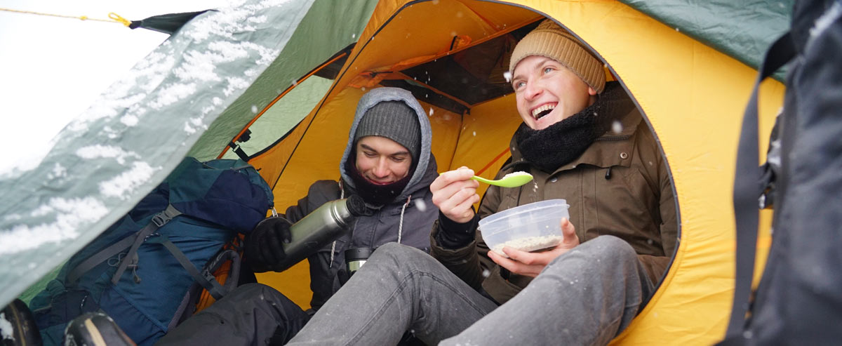 Two winter campers sitting in a tent enjoying warm food and drinks from a thermos