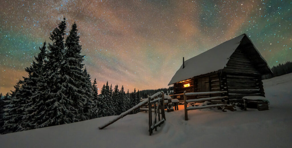 A cozy log cabin with one lit window on the side of a snowy mountain slope and pine tree surroundings 